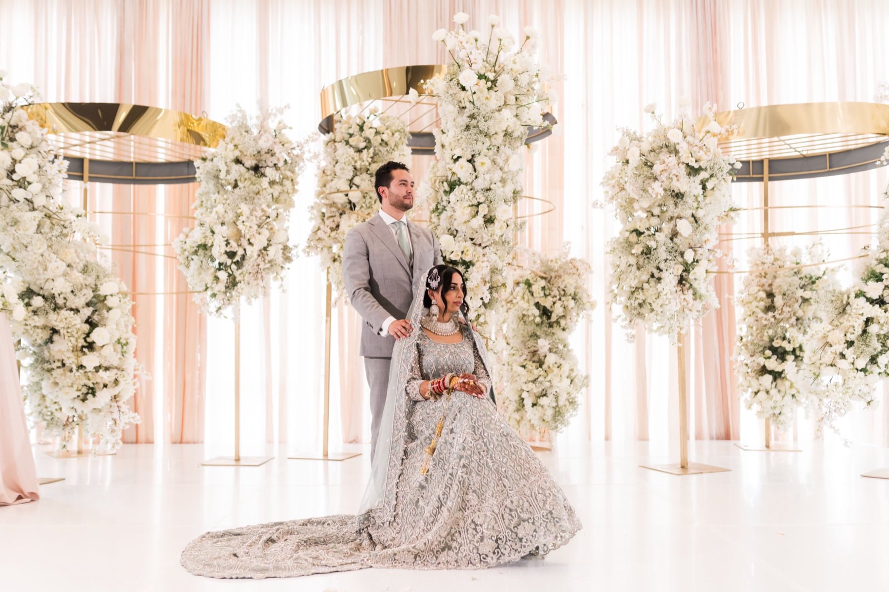 Couple beneath towering white floral installation, bride in silver lehenga