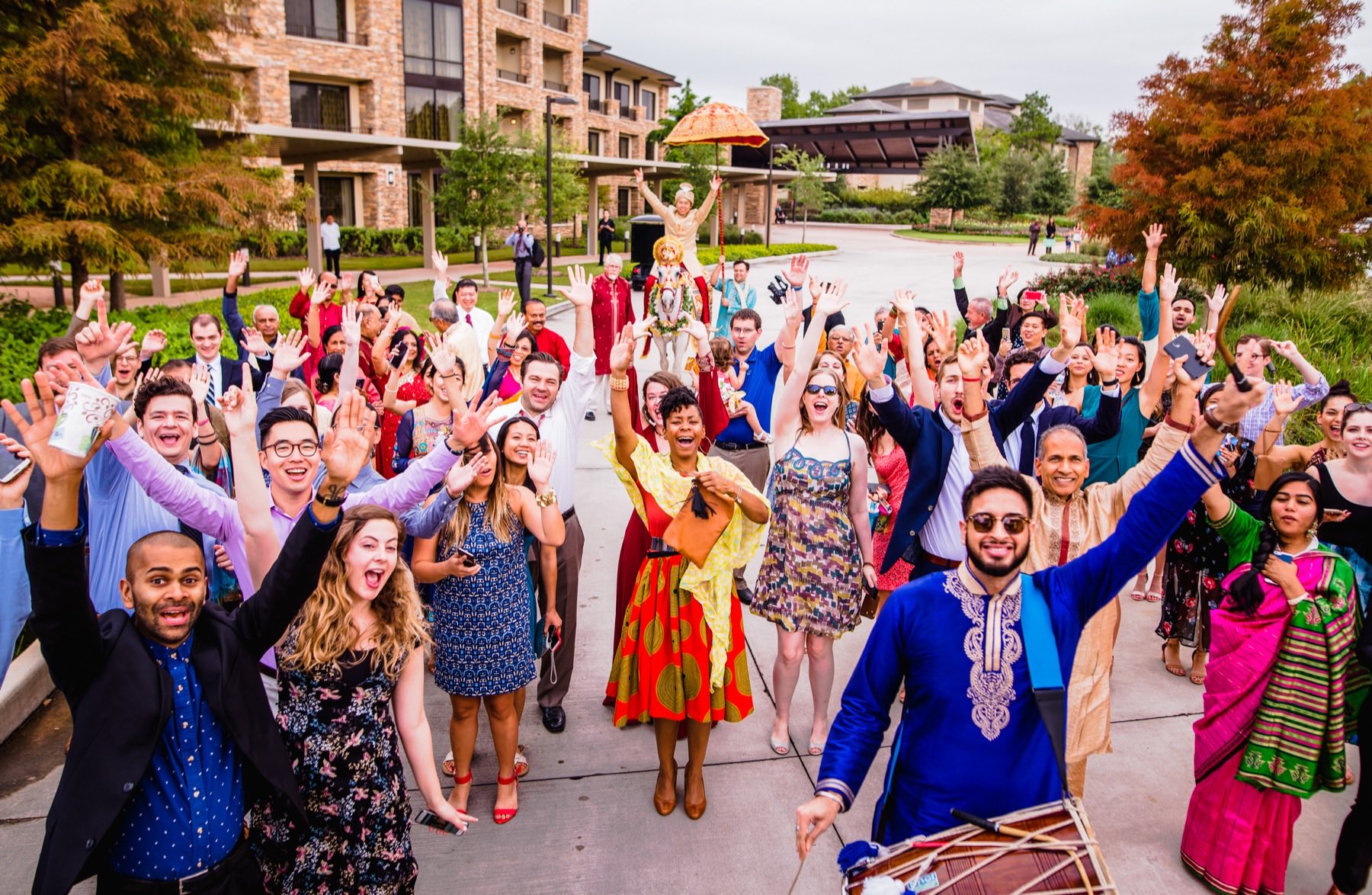Entire multicultural baraat crowd with arms raised, groom on palanquin