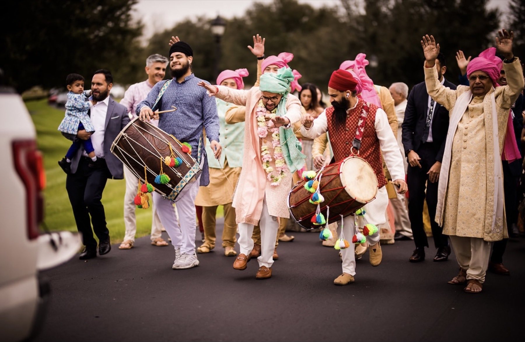 Groom dancing alongside dhol players and guests in pink turbans at baraat procession