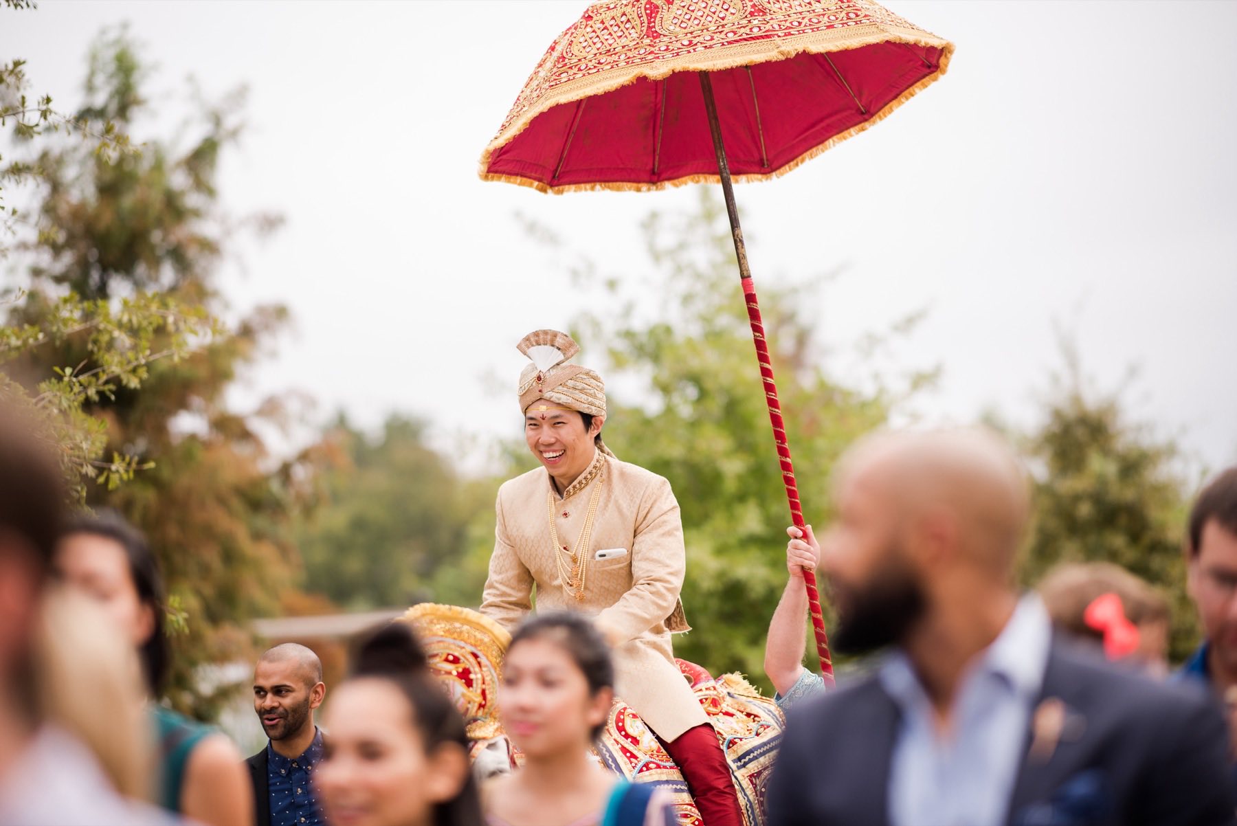 Groom laughing in gold sherwani atop a decorated elephant under a red ceremonial umbrella