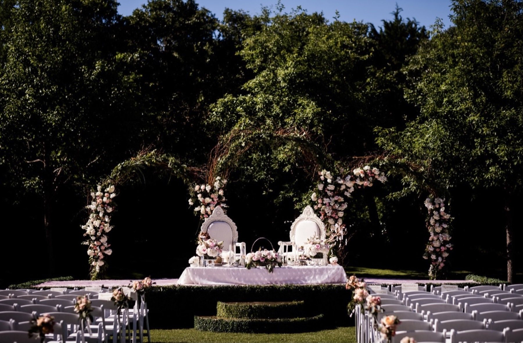 Outdoor ceremony setup with white floral arch and white chairs on lush grounds