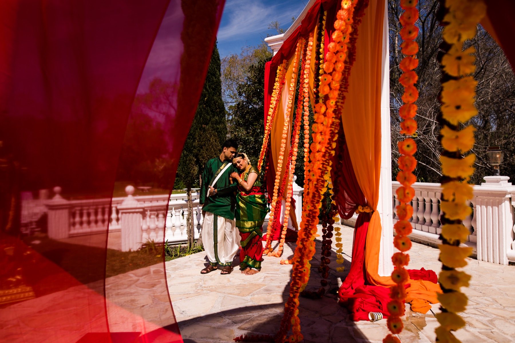 Couple standing together beneath orange marigold mandap framed by red and orange fabric
