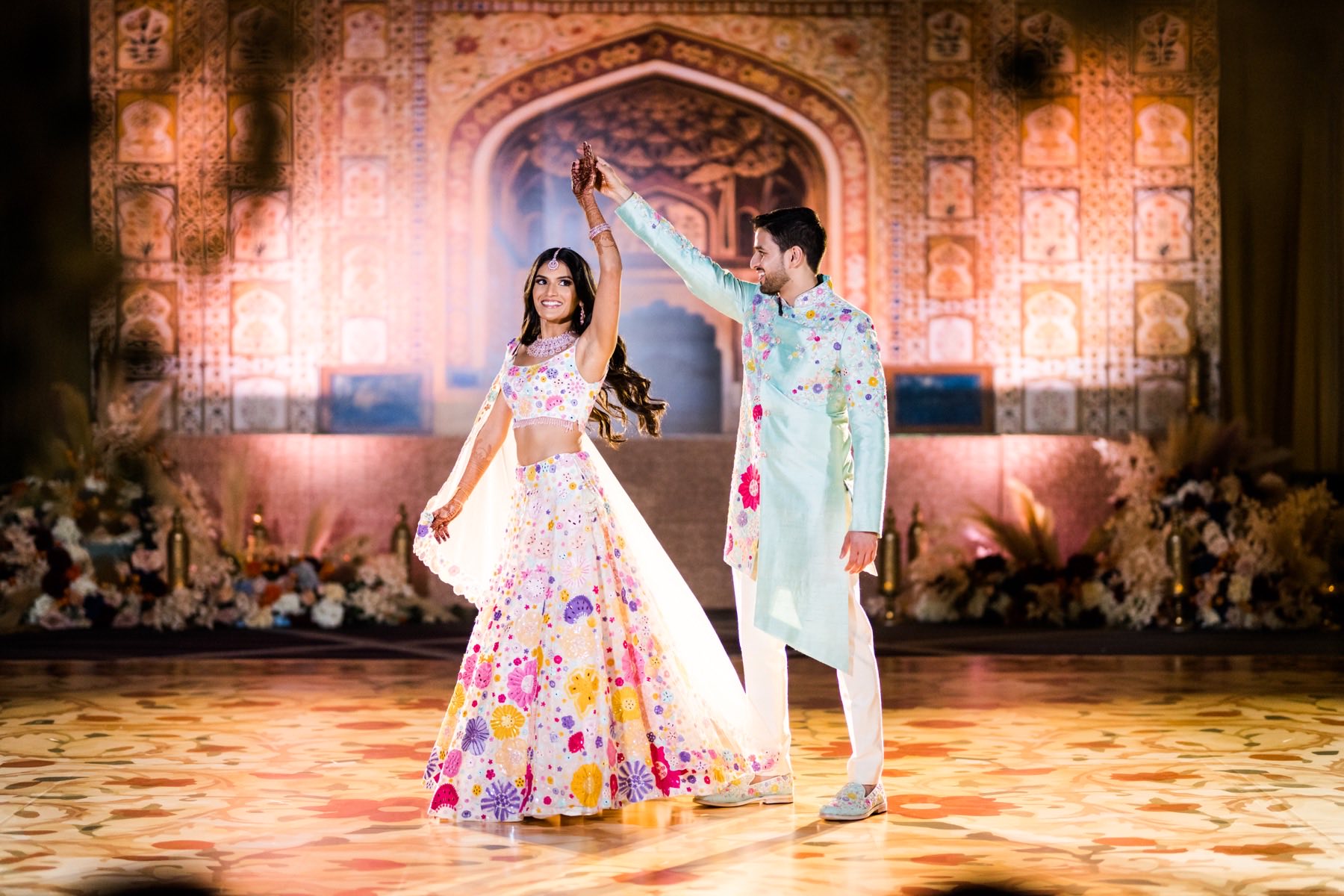Couple twirling together at sangeet, bride in white and floral lehenga on painted floor